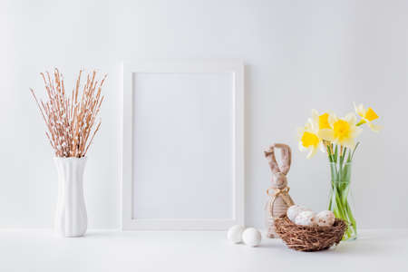 Home interior with easter decor. Mockup with a white frame and yellow daffodils in a glass vase on a light backgroundの写真素材