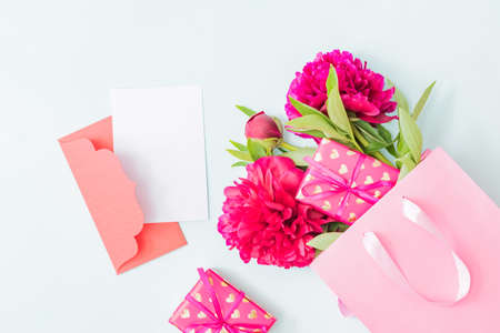 Mockup white greeting card end envelope with red peonies in a pink paper bag and gift box on a light...