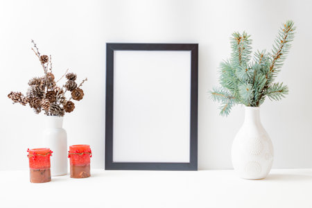 Christmas home interior with decor elements. Mockup with a black frame, spruce branches in a vase, candle, cones on a light backgroundの写真素材