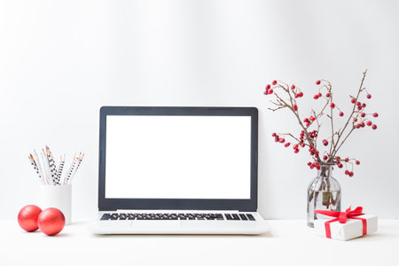 Office workspace with laptop mockup screen, branches with red berries in a glass vase, gift box, supplies on a light background. Christmas home interior or office backgroundの写真素材