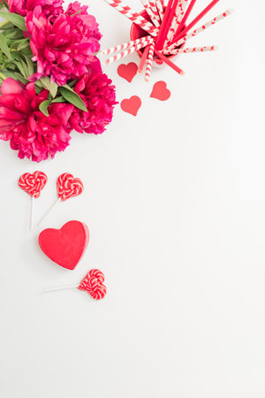 Flat lay valentines day frame with red peonies, lollipops and red hearts on a white background. Valentines day banner with copy spaceの写真素材