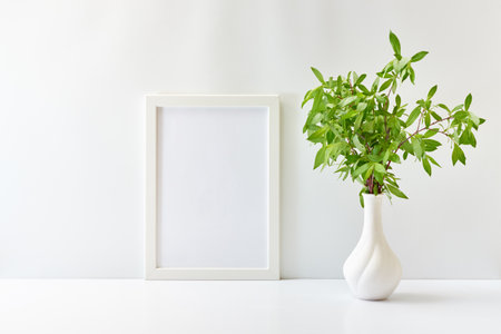 Home interior decor elements. Mockup with a white frame and branches with green leaves in a vase on a white tableの写真素材