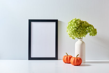 Mockup with a black frame and green flowers in a vase, pumpkins on a light background. Empty poster frame mockup for presentation design, text, letteringの写真素材