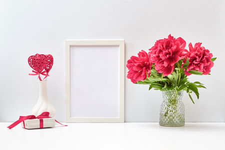 Mockup with a white frame and red peonies in a vase, red heart and gift box on a white table. Empty poster frame mockup for presentation, design, lettering. Valentines Day, Happy Women's Day conceptの写真素材