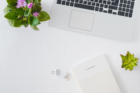 Flat lay home office desktop with laptop, flowers in a pot, notebook, office supplies on a light...