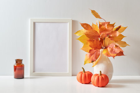 Mockup with a white frame and colorful autumn leaves in a vase on a light background. Empty poster frame mockup for presentation design, text, letteringの写真素材