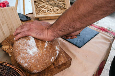 closeup of a man cutting rustic bread with a serrated knife on a wooden board outdoors.の写真素材