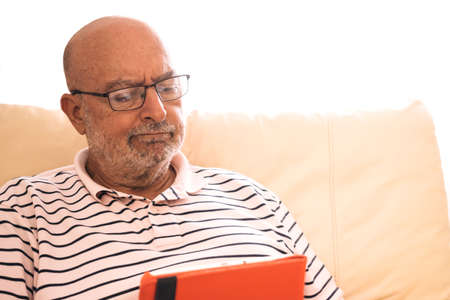 Senior man with glasses looking a digital tablet with angry face at home sitting on the sofa