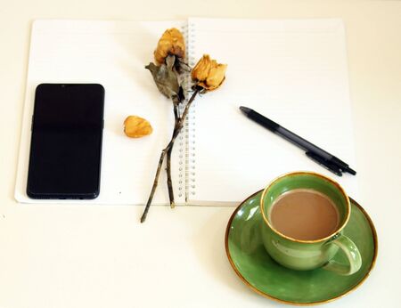 Conceptual photo notebook with pen, classic dried of two roses, black smartphone and a cup of coffee. White notebook and white background.の写真素材