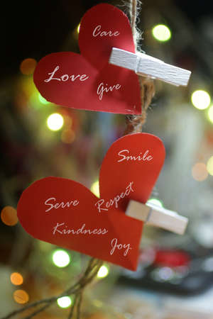 Two love hearts shaped on a string. Red paper with love symbol shape hanging on rope with white wooden clips and positive kindness words. On background of colorful bokeh lights.の写真素材