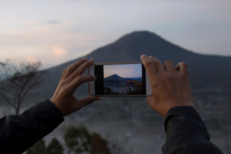 Person with phone camera capturing morning mountain view at sunrise. Smartphone photographer. Person taking Mount Batur picture, Kintamani, Bali, Indonesiaの写真素材
