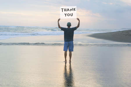 Thank you. Man standing alone with arm raised holding thank you sign on the beach background. Grateful concept.の写真素材