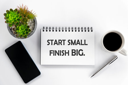 Positive motivational quote on notepaper book - Start small. Finish big. Flat lay concept with plant, phone, pen, and a cup of morning coffee on white desk background.の写真素材