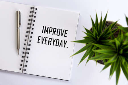 Improve everyday. Single creative word on a notepaper book. Flat lay concept with open book page, pen and green plant on white table background. Reminder for updating business progress and contents.の写真素材
