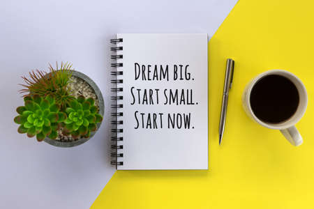 Business motivational quote on a notepaper book - Dream big. Start small. Start now. Flat lay concept with white spiral book, silver pen and green plant on pot on white and yellow table background.の写真素材