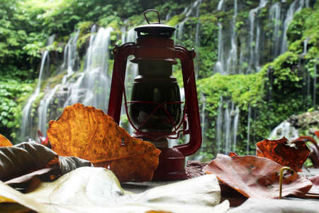 Old classic oil lantern with autumn leaves on blurry background of waterfall in the forest.の写真素材