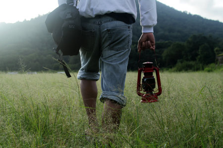 Adventure man walking alone from behind on grass holding oil lantern in hand toward the mountain.の写真素材