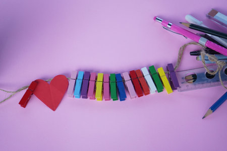 Red heart with colorful wooden clips on rope and pencil pens on soft pink and purple background....