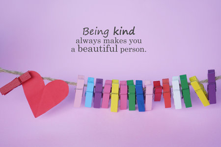 Inspirational quote - Being kind always makes you a beautiful person. With colorful wooden clips on rope and red heart on soft pink purple background. Kindness concept. Be kind.の写真素材