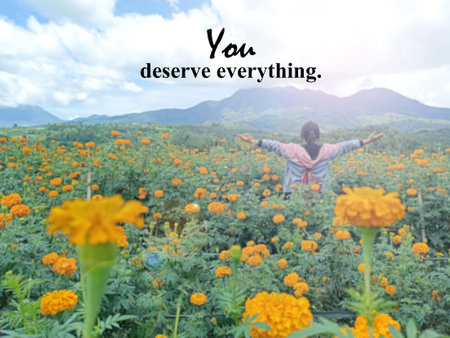 Life inspirational motivational quote - You deserve everything. On background of woman standing with raised hands from behind on marigold flowers garden against the mountain view.の写真素材