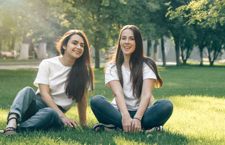 Young beautiful caucasian smiling girls in white t-shirt and jeans sit on the grass. Sexy carefree women posing in the park on a summer day. Girlfriends have fun in sunny weather.の写真素材