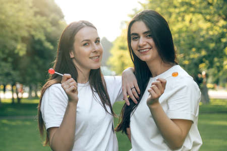 Portrait of two young beautiful European brunettes, smiling girls in a white T-shirt with Chupa Chups caramel in their hands. Sexy Caucasian women posing in the park on a summer day with lollipops in their hands.の写真素材