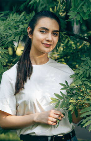 One young beautiful caucasian brunette in a white t-shirt posing in the park on a sunny summer day against the backdrop of green trees. Arabian carefree woman smiling.の写真素材