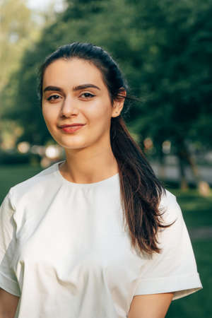 One young beautiful caucasian brunette in a white t-shirt posing in the park on a sunny summer day against the backdrop of green trees. Arabian carefree woman smiling.の写真素材