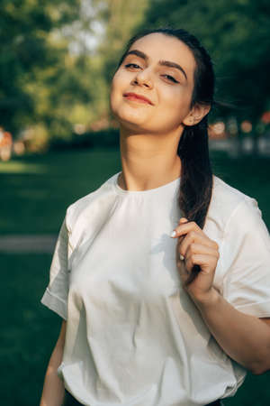 One young beautiful caucasian brunette in a white t-shirt posing in the park on a sunny summer day against the backdrop of green trees. Arabian carefree woman smiling.の写真素材