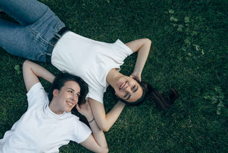 Two young beautiful Caucasian smiling brunettes in a white T-shirt lie on the green grass, laugh and dream. Carefree women have fun chatting on a summer day on the lawn.の写真素材