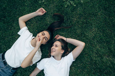 Two young beautiful Caucasian smiling brunettes in a white T-shirt lie on the green grass and laugh. Carefree women have fun chatting on a summer day on the lawn.の写真素材