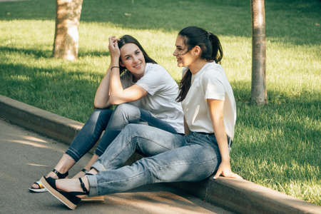 Two young beautiful smiling European girls students in a white t-shirt are sitting on the border in the park on a summer sunny day. Carefree women socialize and talkの写真素材