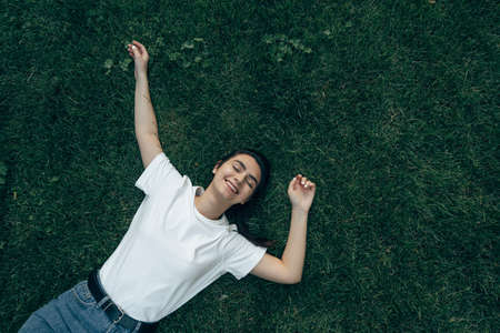 portrait one young beautiful european brunette with dark eyes in a white t-shirt lies on the grass and looks to the side. Caucasian woman lying on the lawn on a summer day. Close-up. Place for text.の写真素材