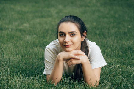 portrait one young beautiful european brunette with dark eyes in a white t-shirt lies on the grass and looks to the side. Caucasian woman lying on the lawn on a summer day. Close-up. Place for text.の写真素材