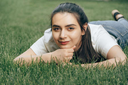 portrait one young beautiful european brunette with dark eyes in a white t-shirt lies on the grass and looks to the side. Caucasian woman lying on the lawn on a summer day. Close-up. Place for text.の写真素材