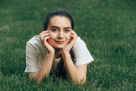 portrait one young beautiful european brunette with dark eyes in a white t-shirt lies on the grass holding her head with her hands and smiling. Caucasian woman lying on the lawn on a summer day. Place for text.の写真素材
