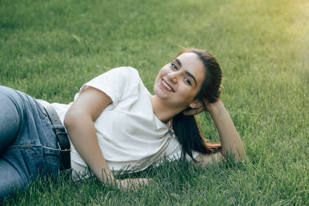 One young beautiful European brunette with dark eyes in a white T-shirt is lying on the grass holding her head and smiling. Caucasian woman laughing while lying on the lawn on a summer day. Place for text.の写真素材