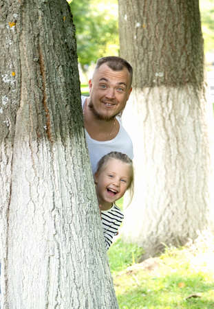 Portrait of a Caucasian little girl looking out from behind a tree with her dad in a park on a sunny day. Father's day. Family weekend in the summer.の写真素材
