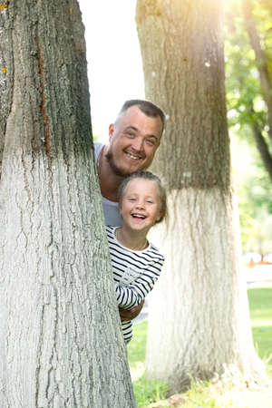 Portrait of a Caucasian little girl 5-6 years old peeping out from behind a tree with her dad in a park on a sunny day. Father's day. Family conceptの写真素材
