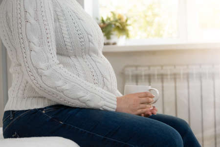 Close-up of a pregnant caucasian woman with a big belly holding a cup and drinking tea at home, sitting on the sofa against the background of the window. Waiting conceptの写真素材