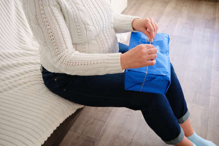 close-up of a young pregnant woman dressed in a white sweater and jeans, opening a blue gift box,...