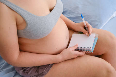 Close-up of a pregnant woman making a packing list for a maternity hospital at home. The expectant mother is engaged in planning, writes shopping lists in a notebook while sitting on the bedの写真素材