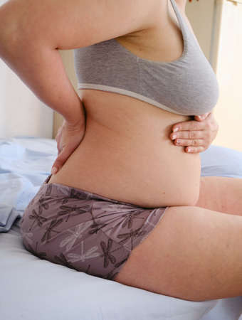 Back pain of a pregnant woman sitting on a bed in the bedroom, holding her lower back and stomach....