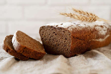 a loaf of home-made dark fragrant fresh bread with sliced slices on a wooden Board against a white brick wall with wheat ears. The concept of rustic home baking.の写真素材