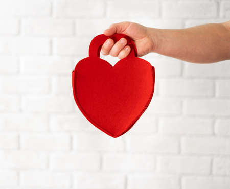 Caucasian man holding a red heart against a white brick wall. Men's hands close up. Valentine's day, a gift to your beloved womanの写真素材