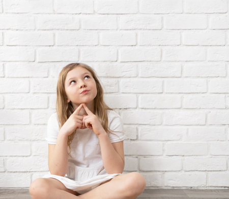 Portrait of a happy child - a little blonde girl 8-9-10 years old, against the background of an empty white brick wall, dressed in rabbit ears, holding hands in the shape of a heart. Medium planの写真素材