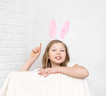A small emotional blonde with pink bunny ears on her head sits looking at the camera holding her finger up, wondering at the idea. The concept of a holiday is Easter, Children's Day.の写真素材