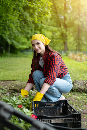 A young woman is an agricultural worker working with flowers in a greenhouse. a girl in yellow gloves, shirt and jeans on the background of a plantation. The concept of natural gardeningの写真素材