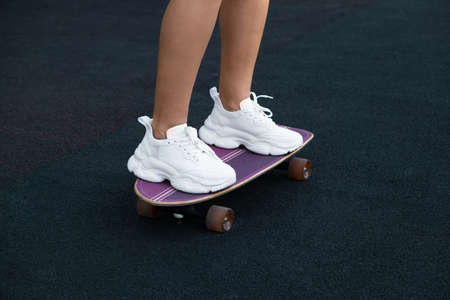 close-up of the female legs of a Caucasian girl, shod in white women's sneakers, standing on a purple skateboard in the park against the background of a treadmillの写真素材