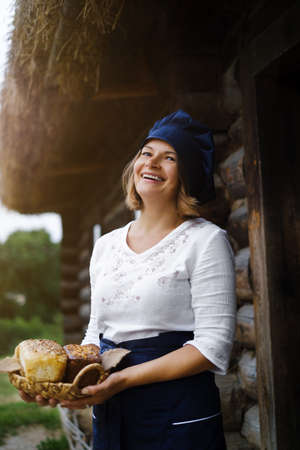 a smiling baker woman with a basket of bread in her hands in a chef's hat and apron against the background of a wooden log village house. Yeast-free wheat and rye bread with sourdough. Eco bakery.の写真素材